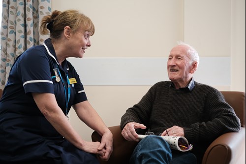 A nurse in a blue uniform is speaking with a patient, an older man who is sitting in a brown chair. They are smiling and chatting.