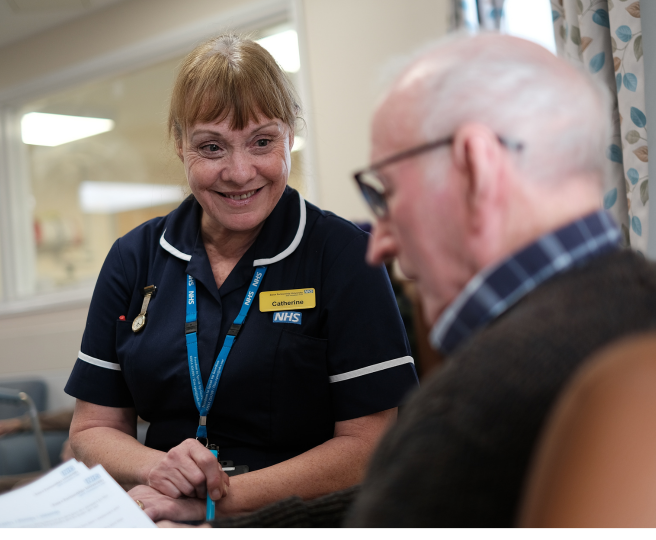 Nurse in a dark blue top with and NHS lanyard speaking to an elderly male patient.