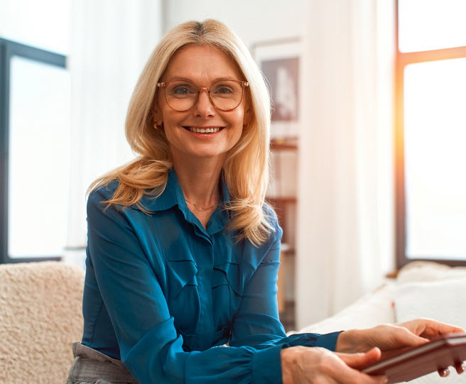 Sitting on a sofa, blonde haired lady, wearing glasses and blue top holding a pad