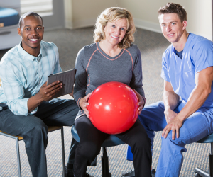 Two men sitting and one lady sitting together. The black man is holding and iPad and the lady is holding a ball. The Caucasian man is wearing a blue healthcare unifrom.