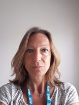 Woman with light brown hair, wearing a patterned white shirt. Has an NHS lanyard around her neck.